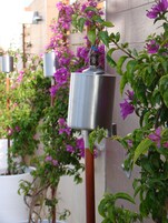 Un dispensador de agua metálico fijado a la pared, con follaje verde y flores púrpuras al fondo.