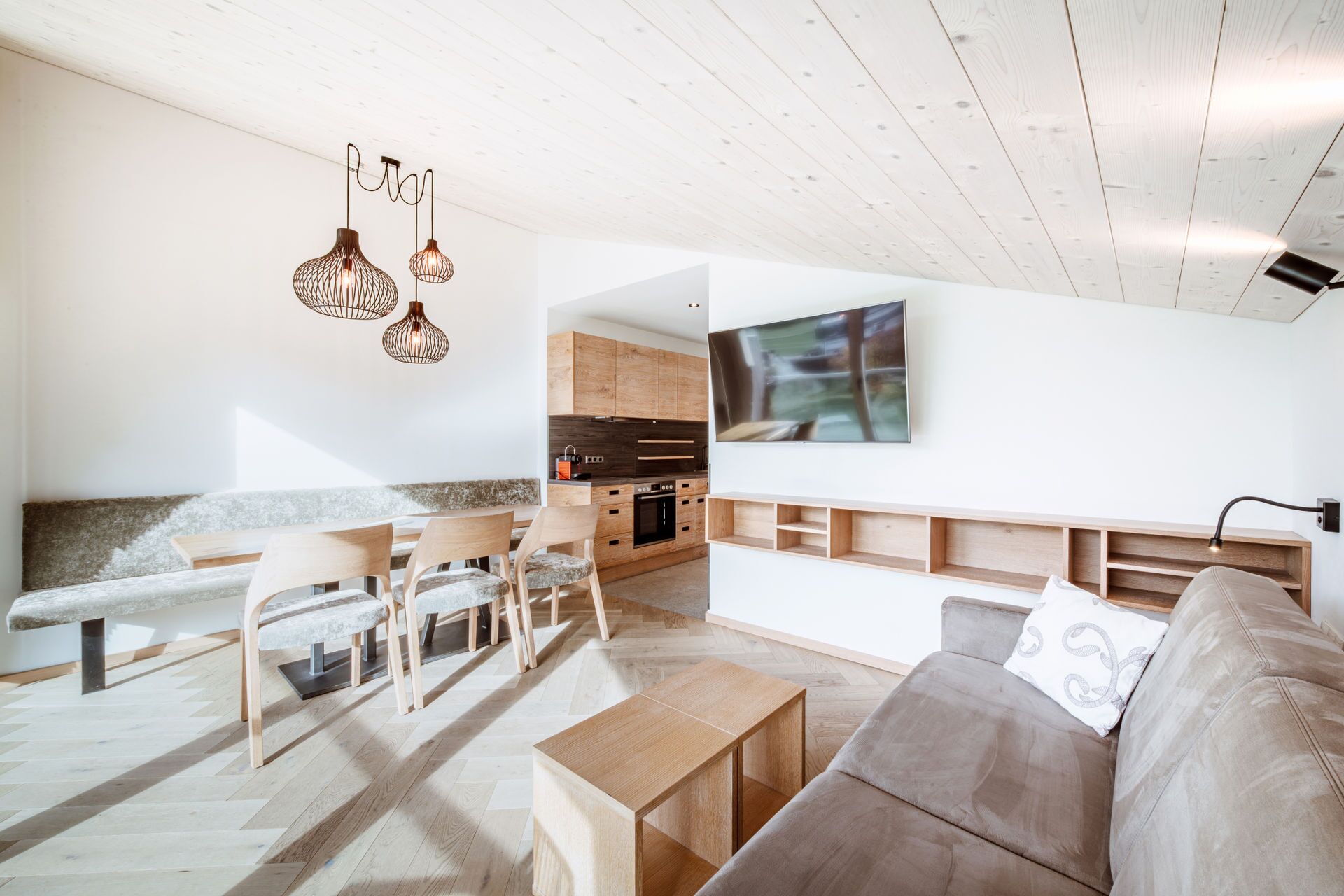 A modern living room with a wooden bench, a sofa, and a dining table. There is a kitchen area with wooden cabinets and a built-in oven.