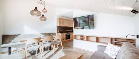Ein modernes Wohnzimmer mit einer hölzernen Bank, einem Sofa und einem Esstisch. Es gibt eine Küchenzeile mit Holzschränken und einem eingebauten Backofen.