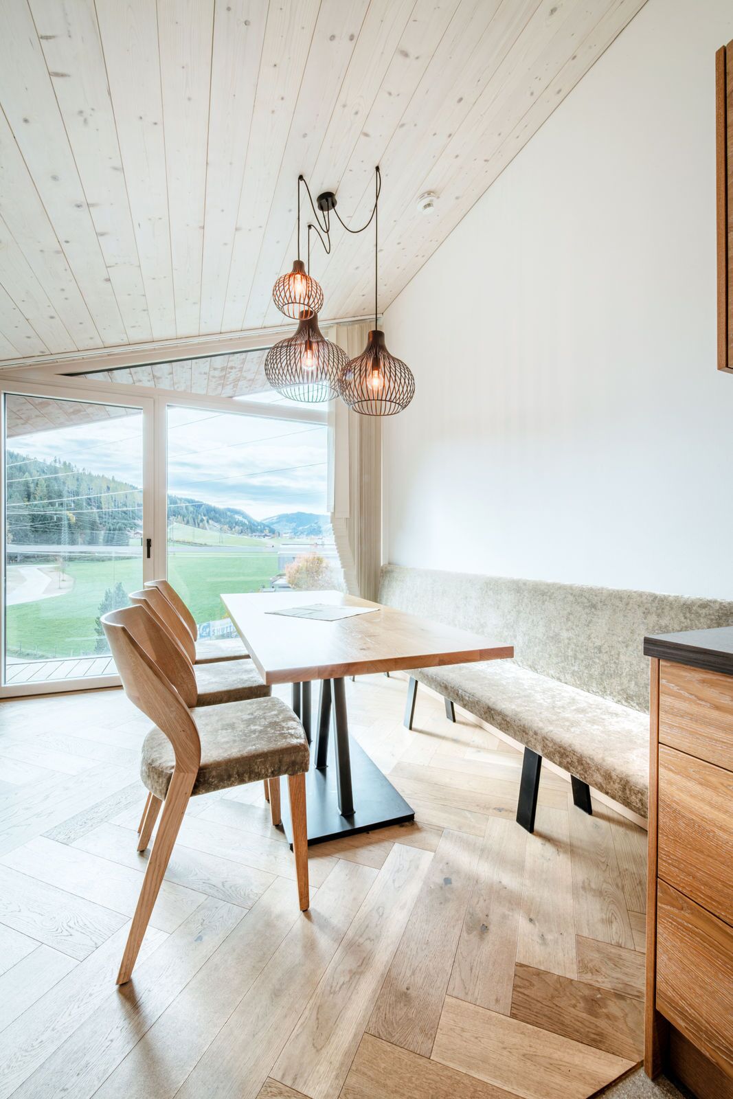 A modern dining area with a wooden table and bench, a view of a green landscape through large windows, and a ceiling with exposed wooden beams.