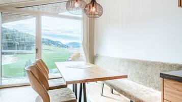 A modern dining area with a wooden table and bench, a view of a green landscape through large windows, and a ceiling with exposed wooden beams.
