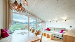 Une pièce spacieuse avec un plafond en bois, une grande fenêtre donnant sur l’extérieur, une table à manger avec des chaises et un canapé avec des coussins.