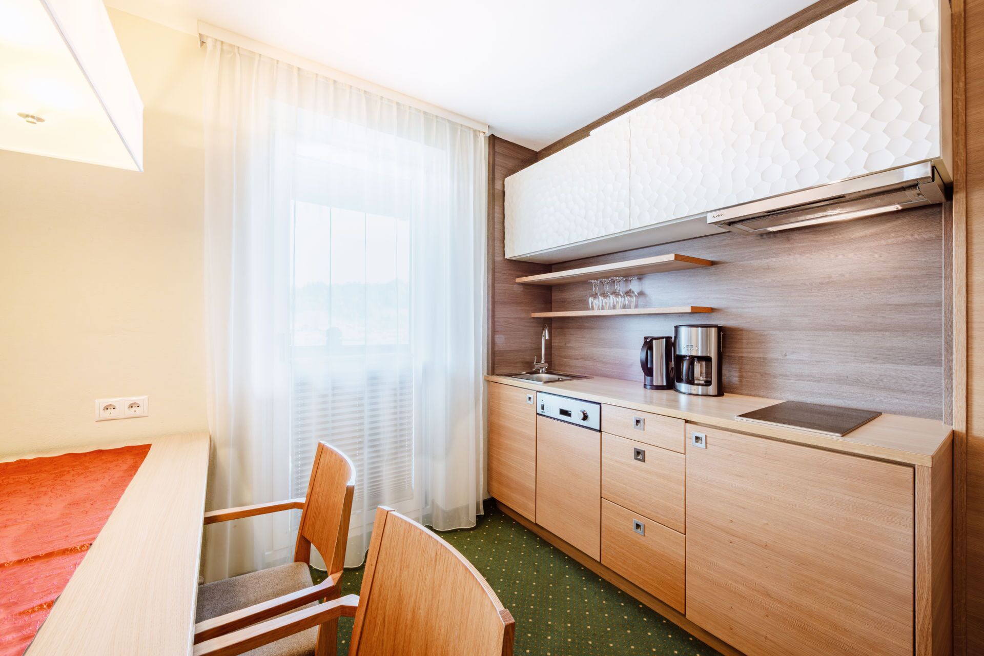Cocina de hotel compacta con lavamanos, cafetera y comedor con una silla.