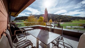 Un balcon avec une table et des chaises, donnant sur un paysage magnifique avec des montagnes et un ciel dégagé.