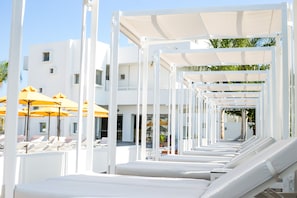 A poolside area with white lounge chairs and yellow umbrellas, a white building in the background, and a clear sky. - Orion Hotel Faliraki (Rhodes)