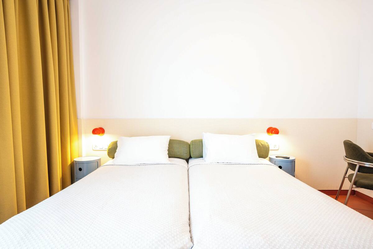 A hotel room with two beds, a nightstand, a chair, and a wall-mounted lamp.