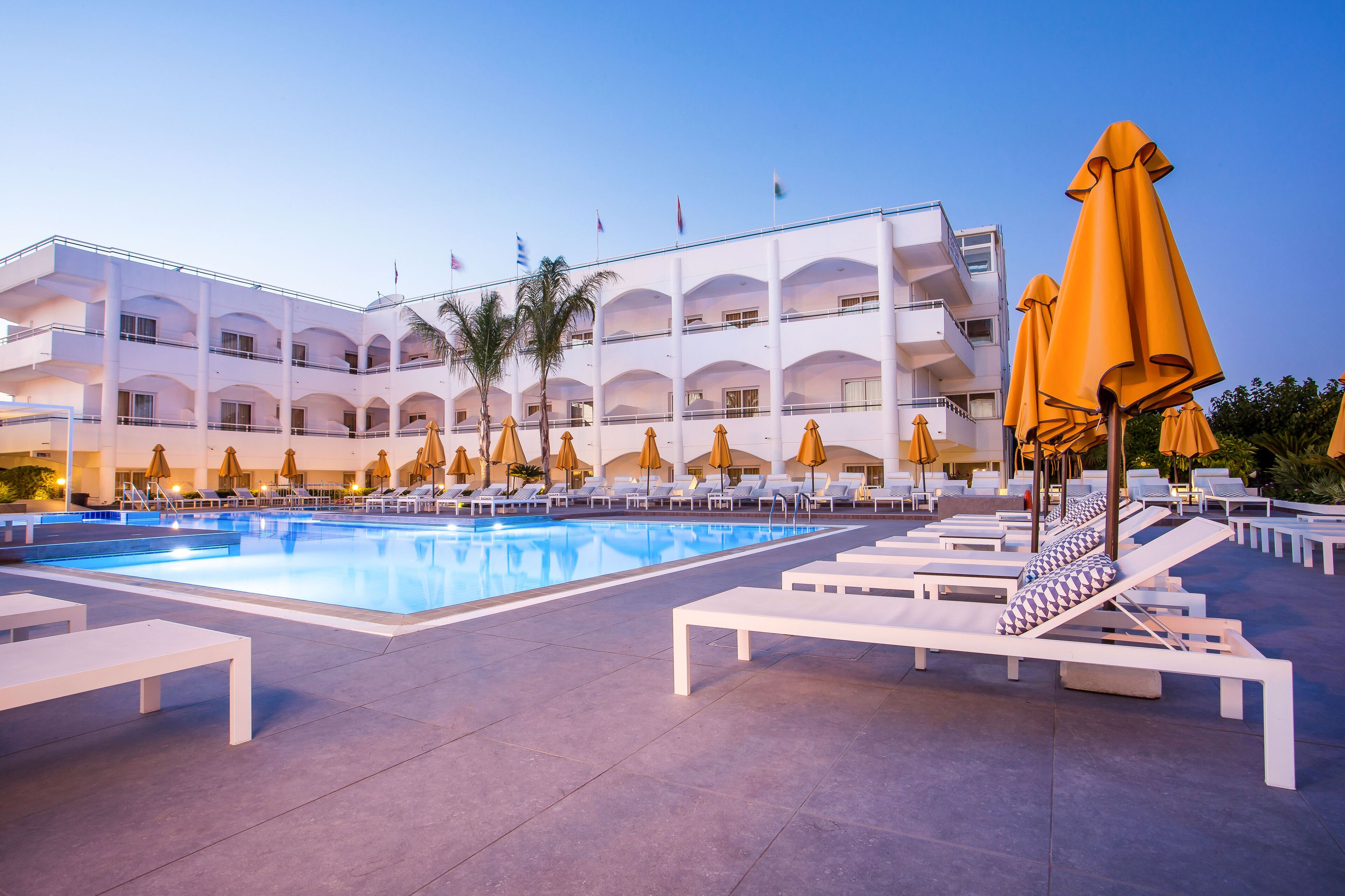 A hotel pool area with sun loungers and umbrellas, a clear blue pool, and a multi-story building with balconies.