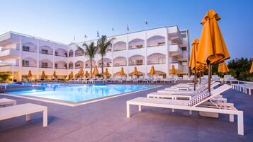 A hotel pool area with sun loungers and umbrellas, a clear blue pool, and a multi-story building with balconies.