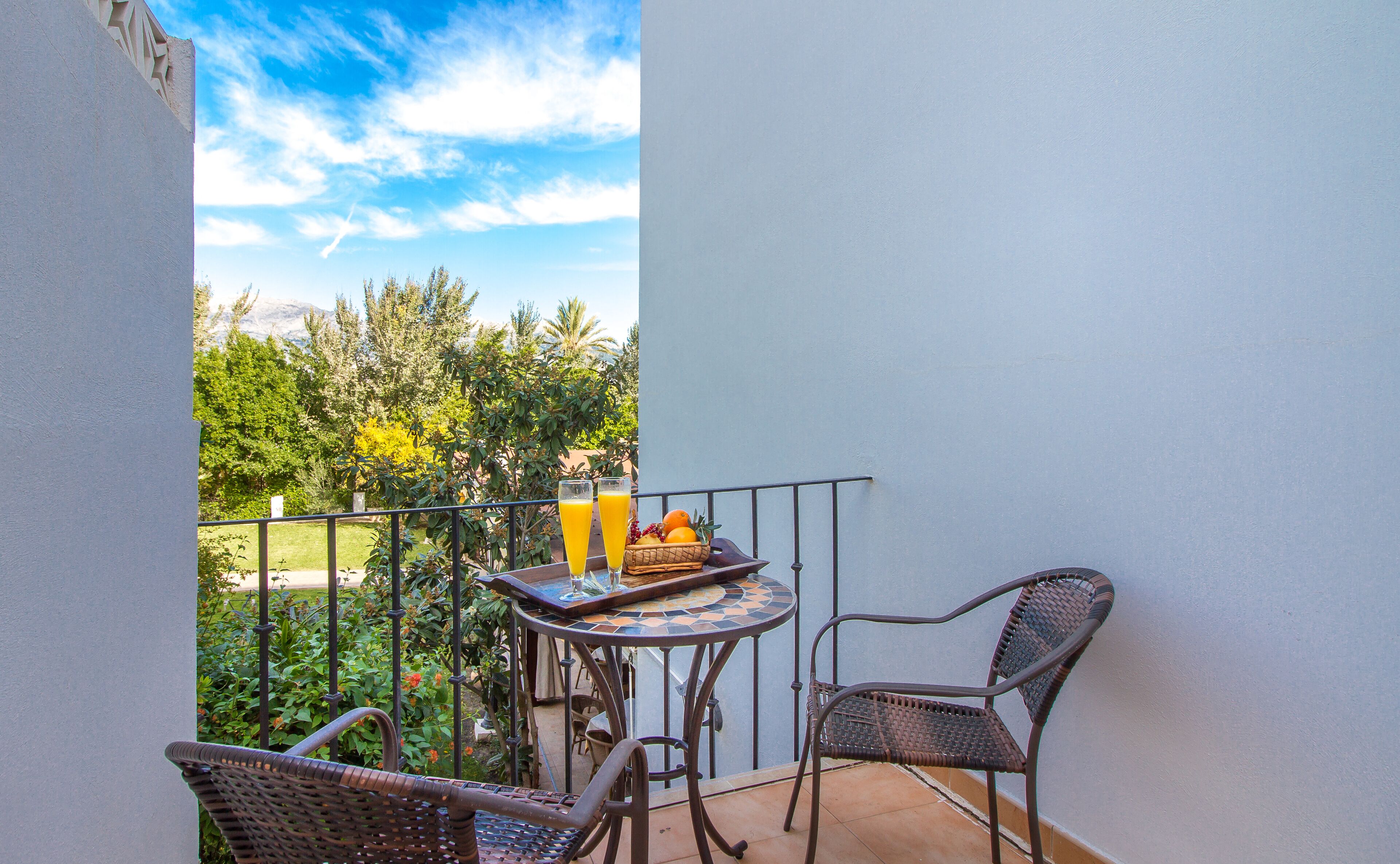 A balcony with a table and two chairs, a fruit basket, and two glasses of orange juice.