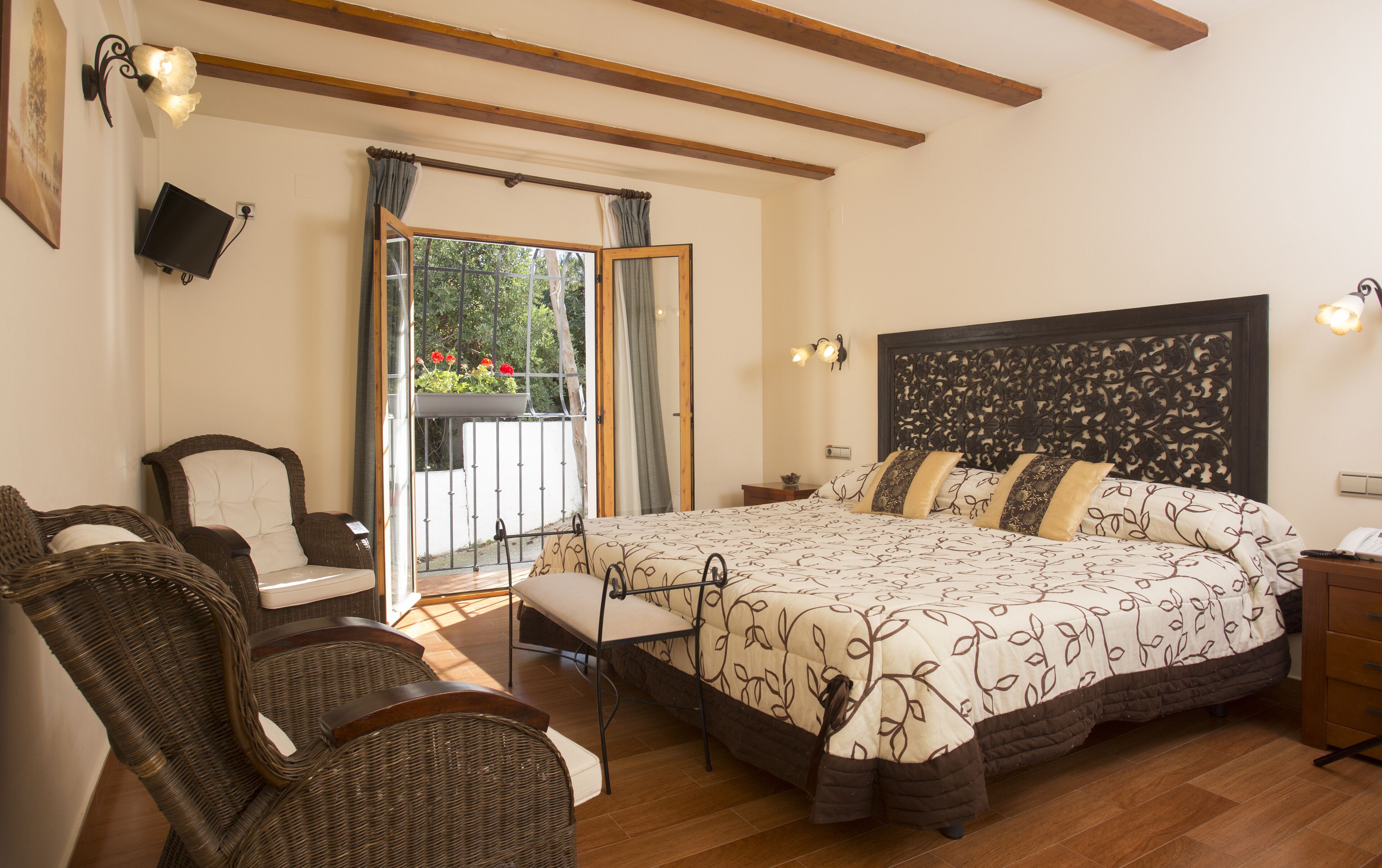 Un dormitorio con una cama grande, cabecera de madera, sillas de mimbre, una mesita y un balcón con flores.