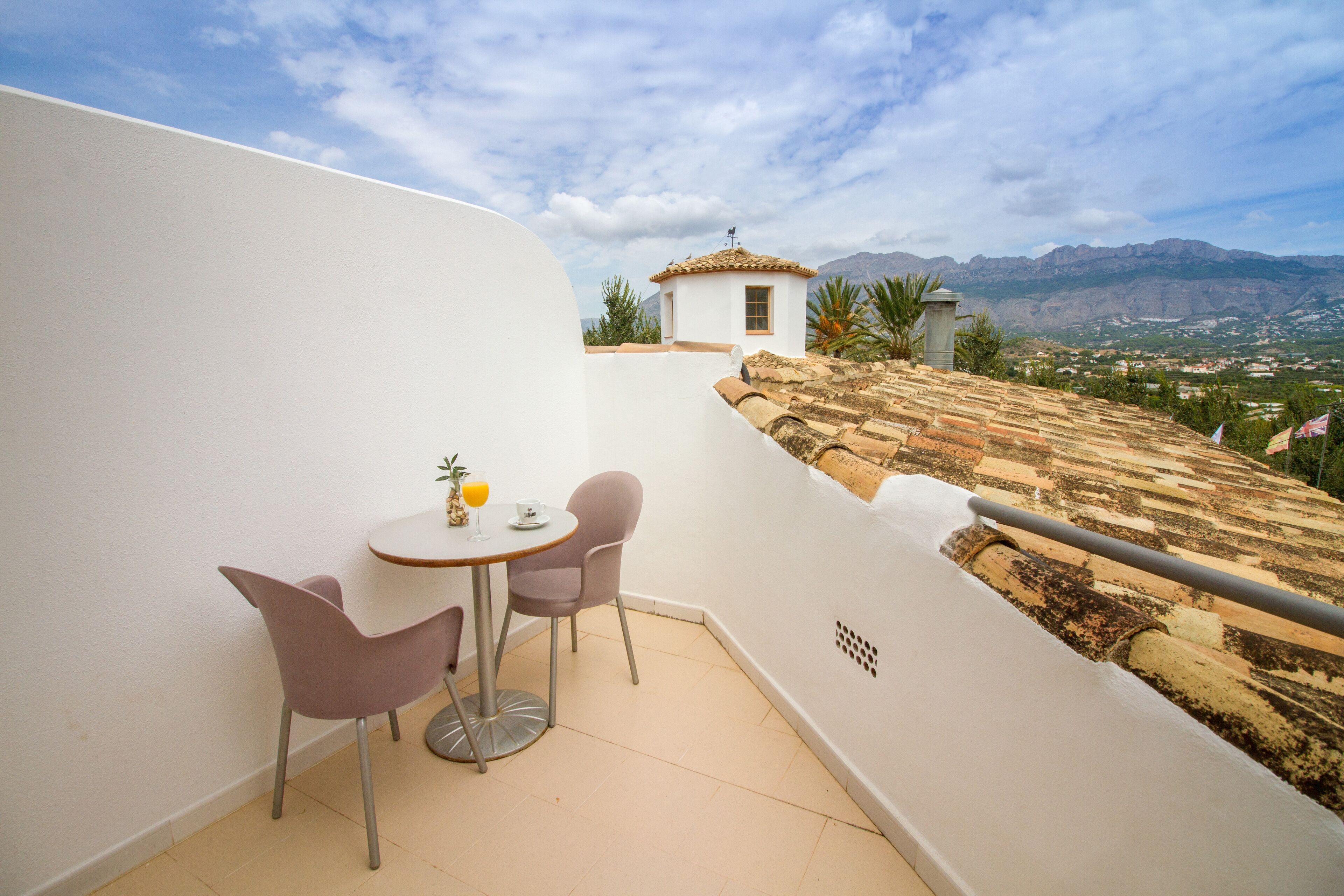 Un balcón con una mesa y sillas, un edificio pequeño al fondo y un paisaje montañoso.