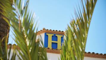 Um edifício com um grande letreiro azul e amarelo em formato de 'H', rodeado por folhas de palmeira.