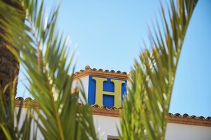Um edifício com um grande letreiro azul e amarelo em formato de 'H', rodeado por folhas de palmeira.