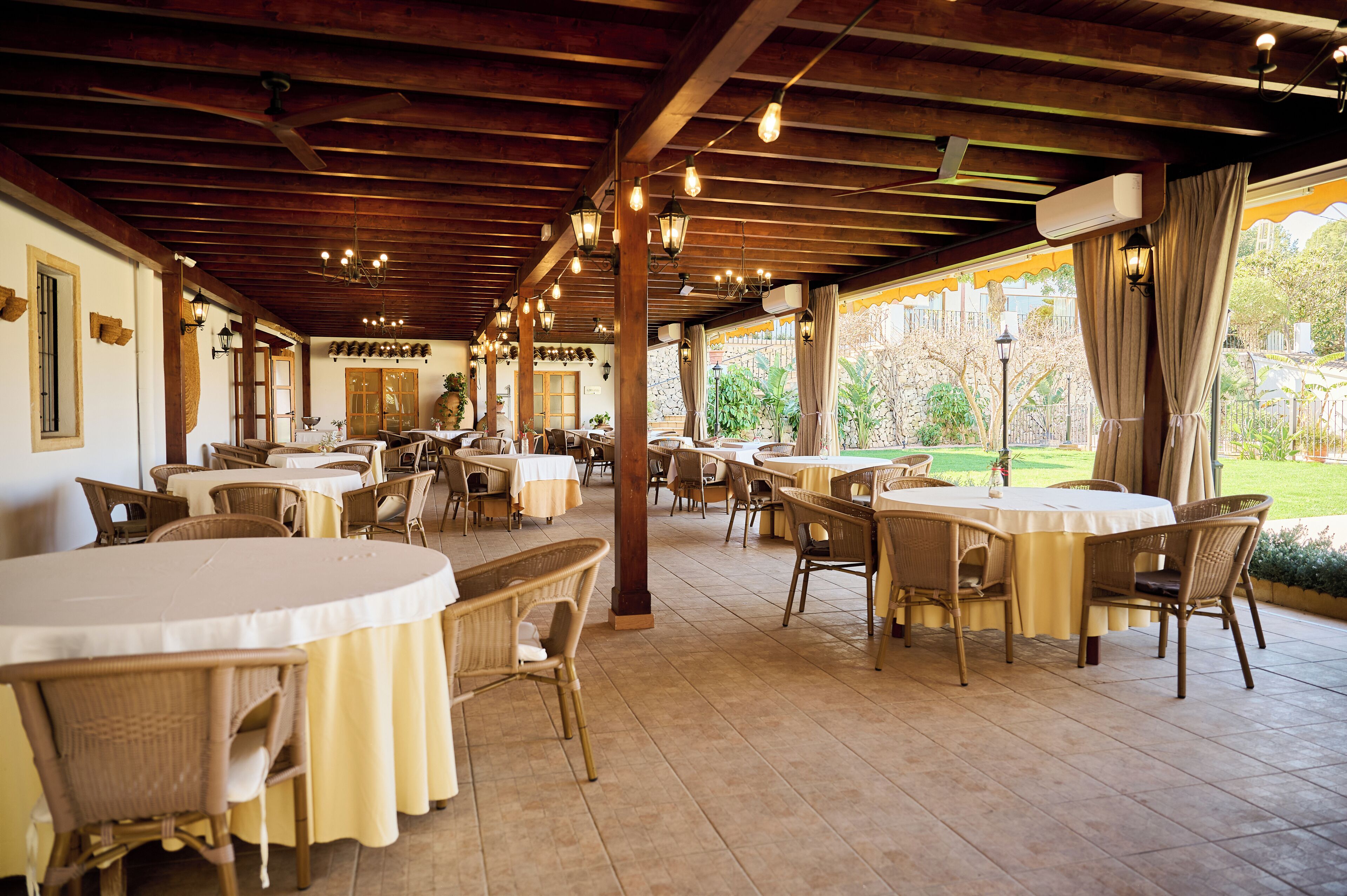 An indoor dining area with round tables and wicker chairs, wooden beams on the ceiling, and large windows overlooking a garden.