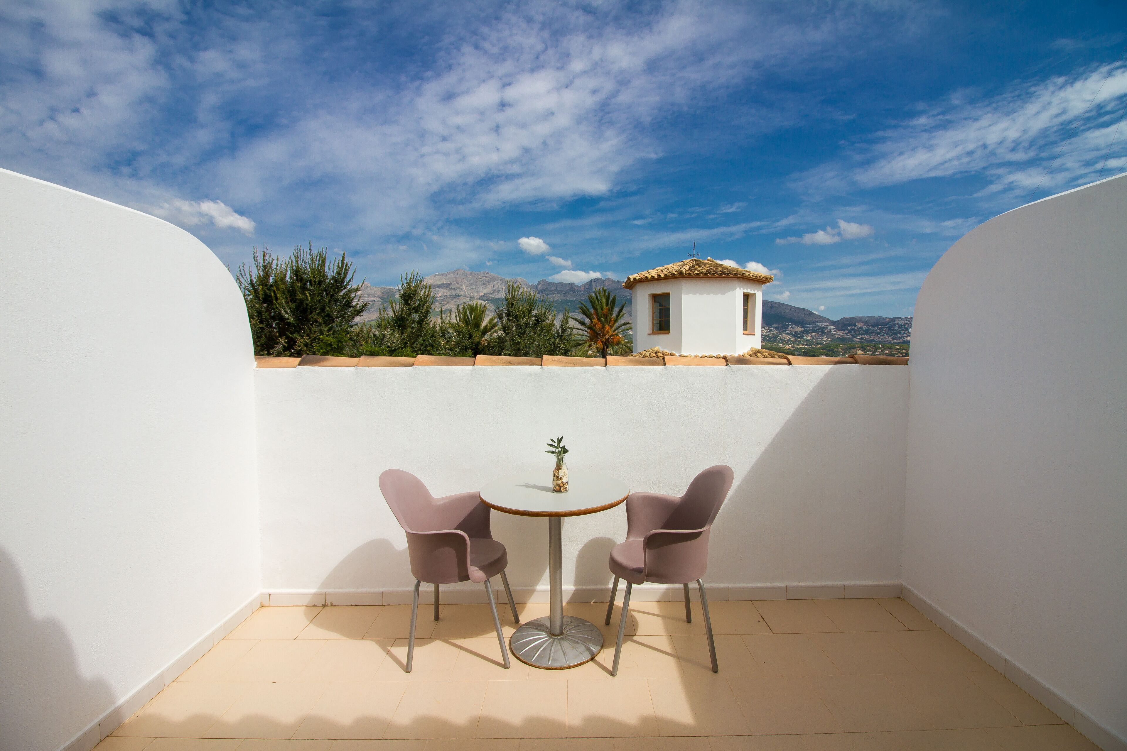 A small balcony with a round table and two chairs, a view of a mountain and a building in the background.