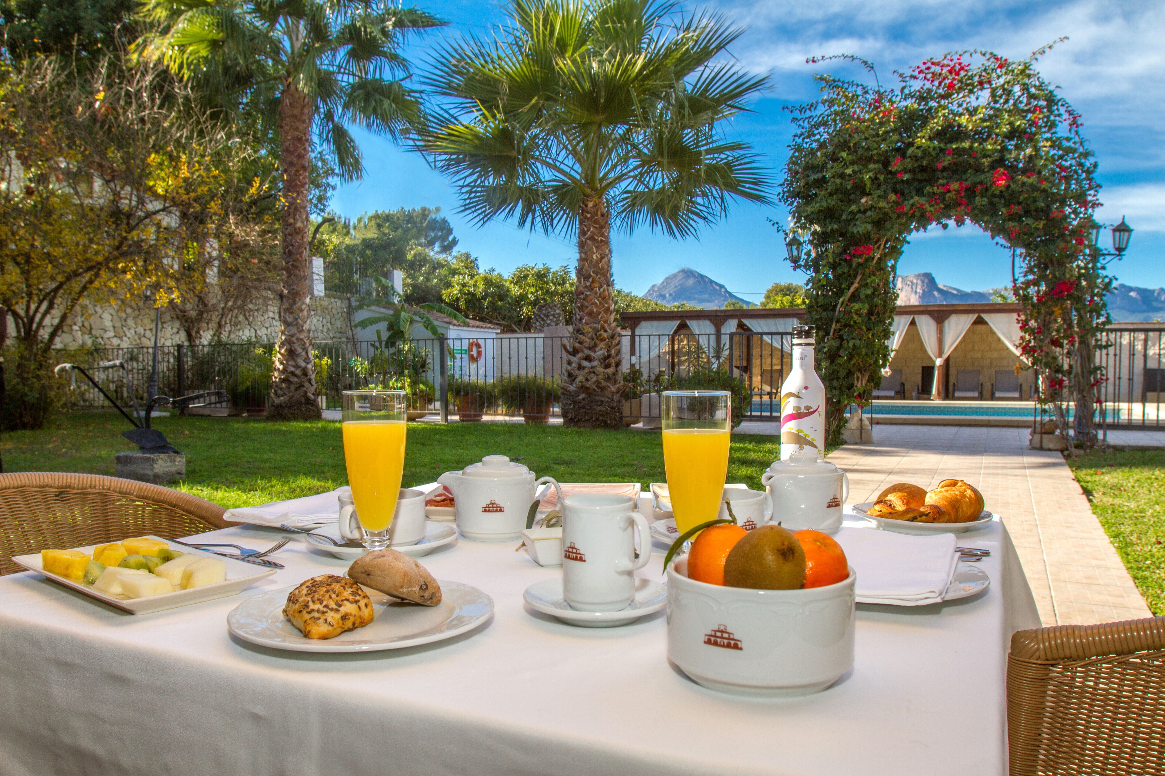 Un desayuno al aire libre con una mesa preparada para dos personas, con platos de fruta, pasteles y una botella de jugo de naranja.