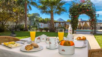 Un desayuno al aire libre con una mesa preparada para dos personas, con platos de fruta, pasteles y una botella de jugo de naranja.