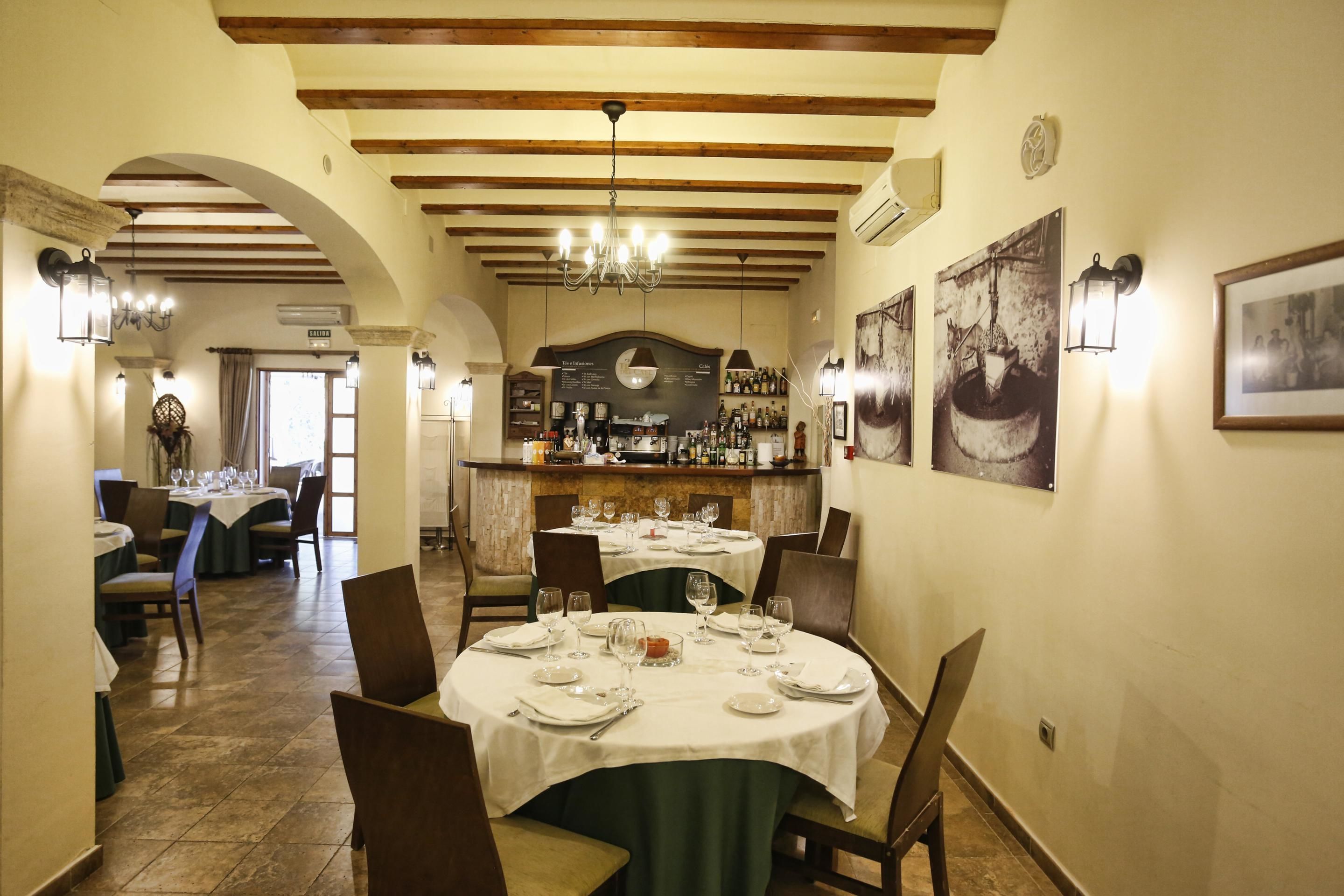 A restaurant with a high ceiling, wooden beams, and a bar area. Tables are set with white tablecloths and chairs.