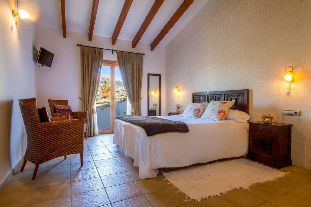Ein Schlafzimmer mit Bett, Sessel, Nachttisch, Fernseher und Blick ins Freie.