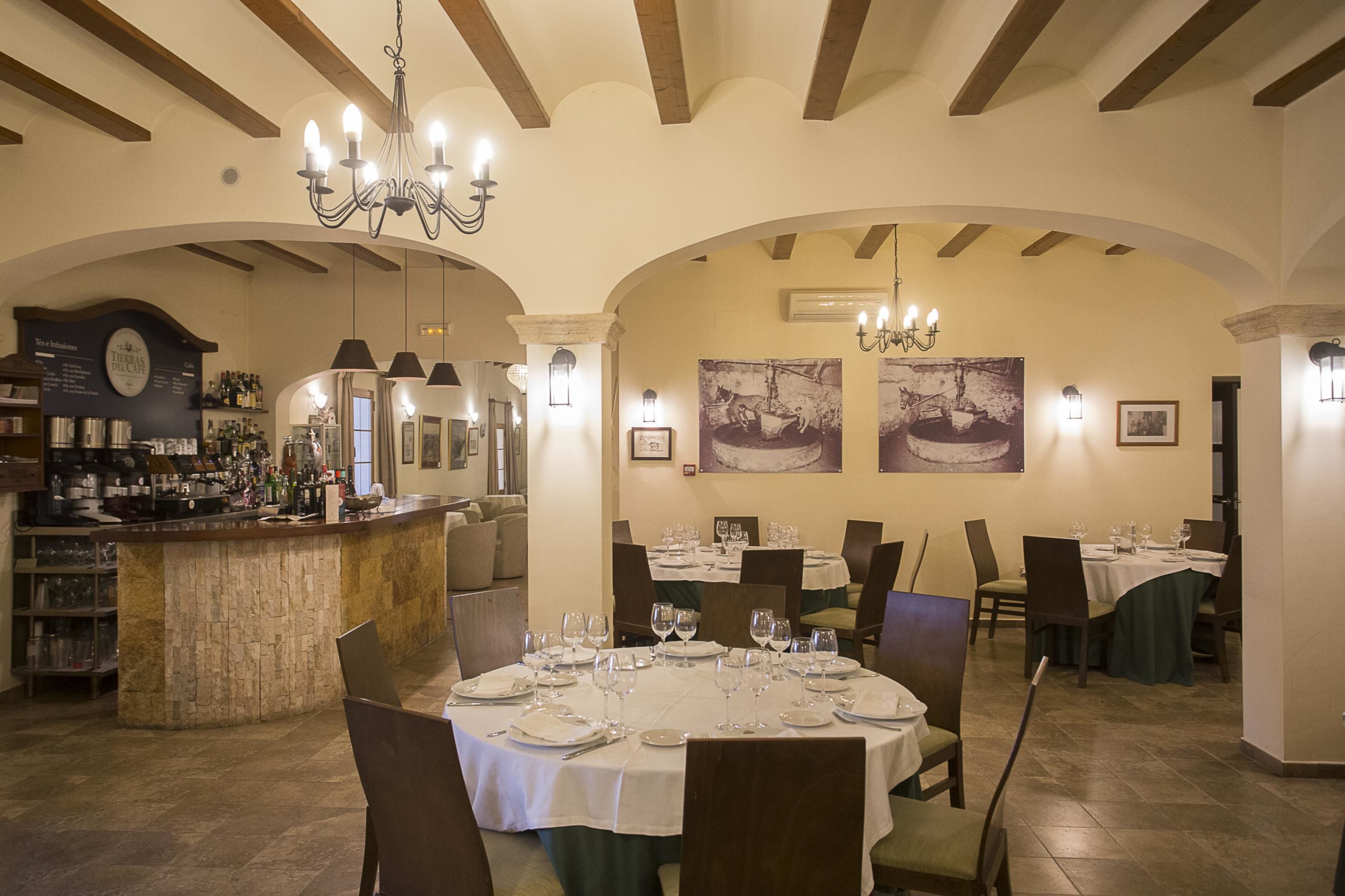Un restaurante con mesas redondas dispuestas para comer, una zona de bar con botellas y un candelabro.