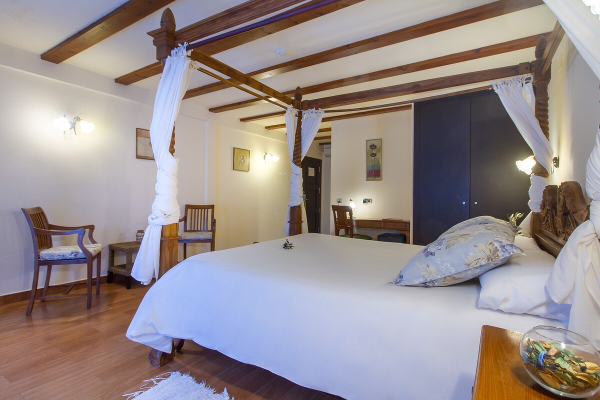 Un dormitorio con cama con dosel, vigas de madera, una silla, una mesita pequeña y una mesita de noche.