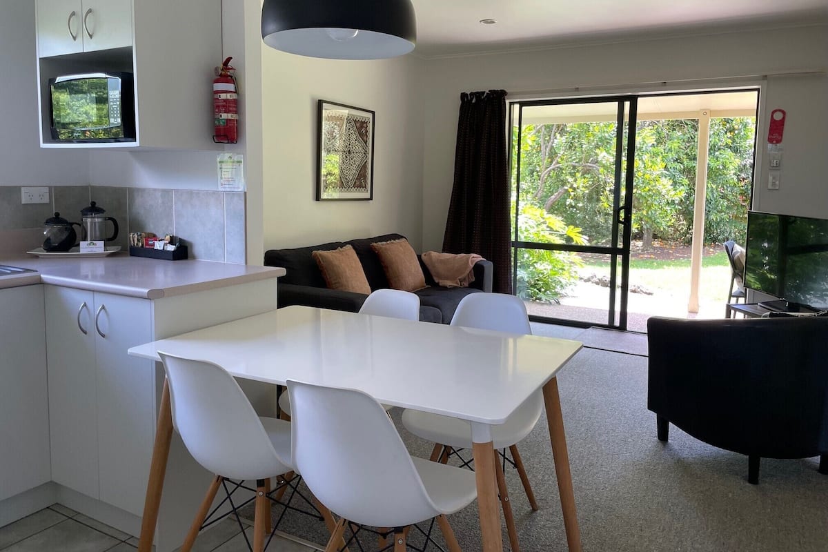 A modern kitchen with a microwave, kettle, and coffee maker. There is a dining area with a white table and chairs. A black sofa and a TV are visible in the living area.