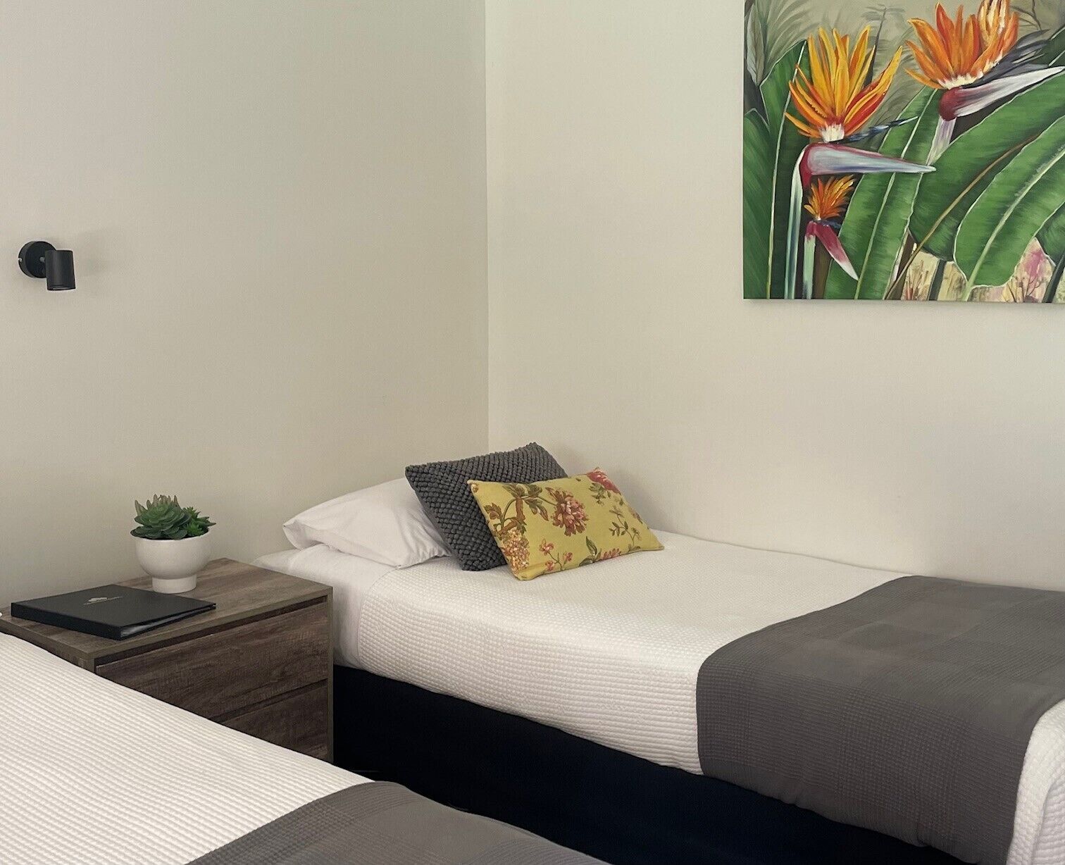 Un dormitorio con una cama individual, una mesita de noche con una planta, una lámpara y un cuadro de flor de paradisíaco.