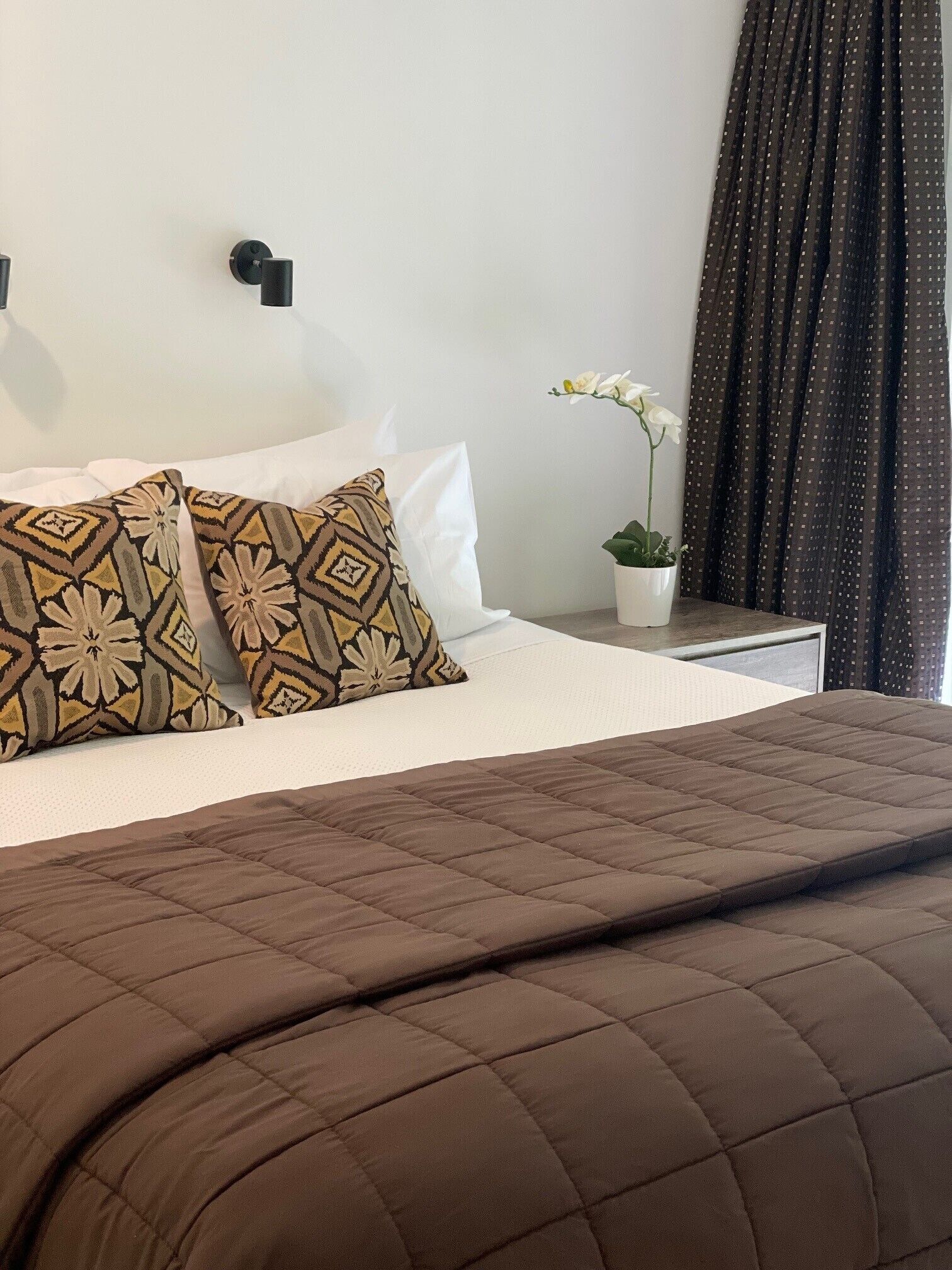 A neatly made bed with a brown quilt, white pillows, and patterned cushions. A potted plant with white flowers is on the nightstand beside the bed.