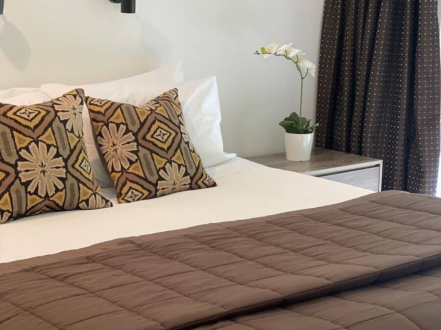 A neatly made bed with a brown quilt, white pillows, and patterned cushions. A potted plant with white flowers is on the nightstand beside the bed.