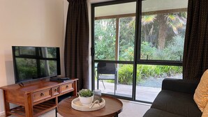 A living room with a TV, wooden furniture, a round coffee table with a plant, and a large window with curtains. - Kauri Park Motel (Kerikeri)