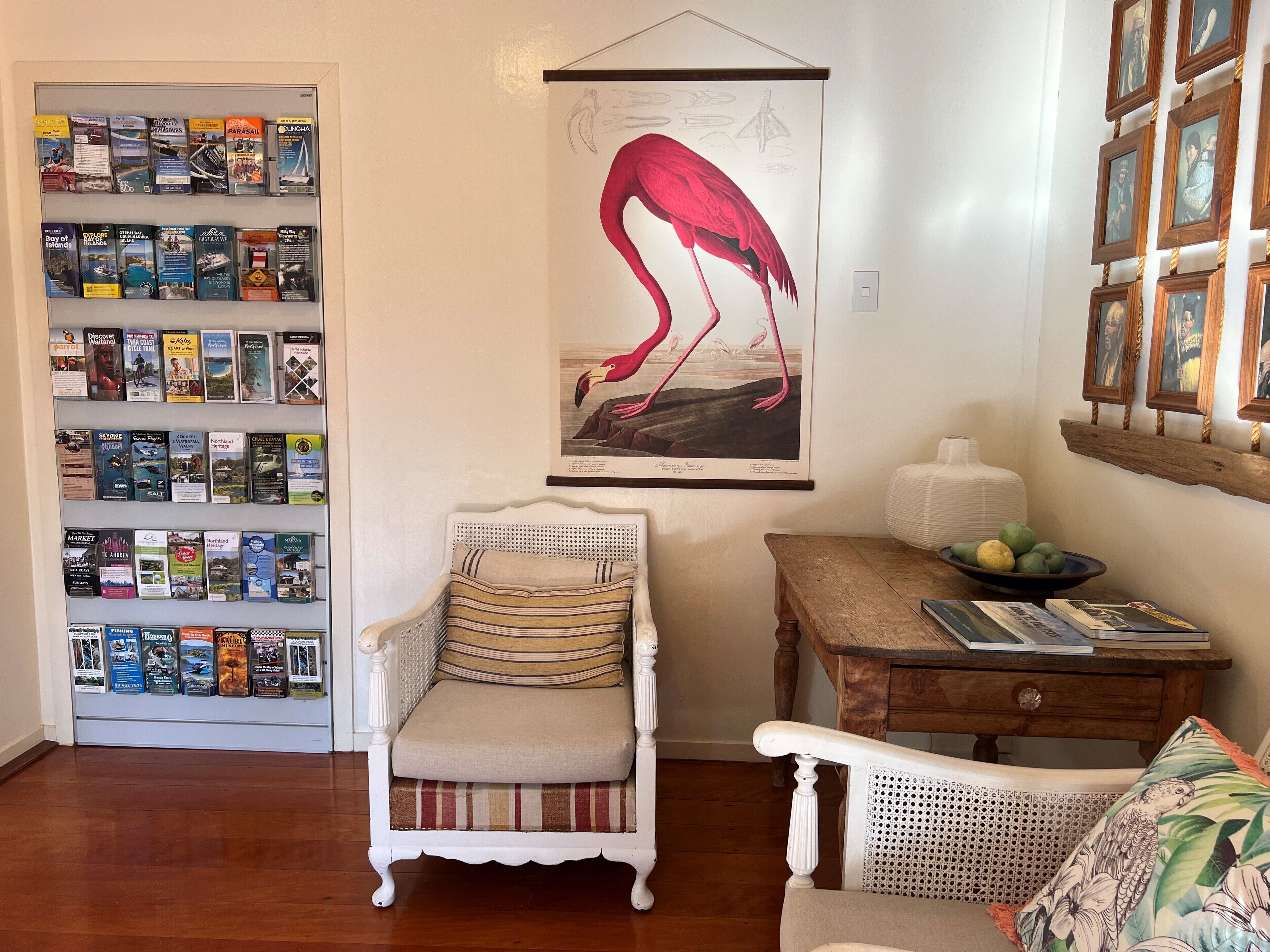 Um quarto com uma estante repleta de livros de viagem, uma ilustração emoldurada de um flamingo, uma cadeira branca, uma mesa de madeira com uma tigela de frutas e uma cadeira de vime com uma almofada florida.