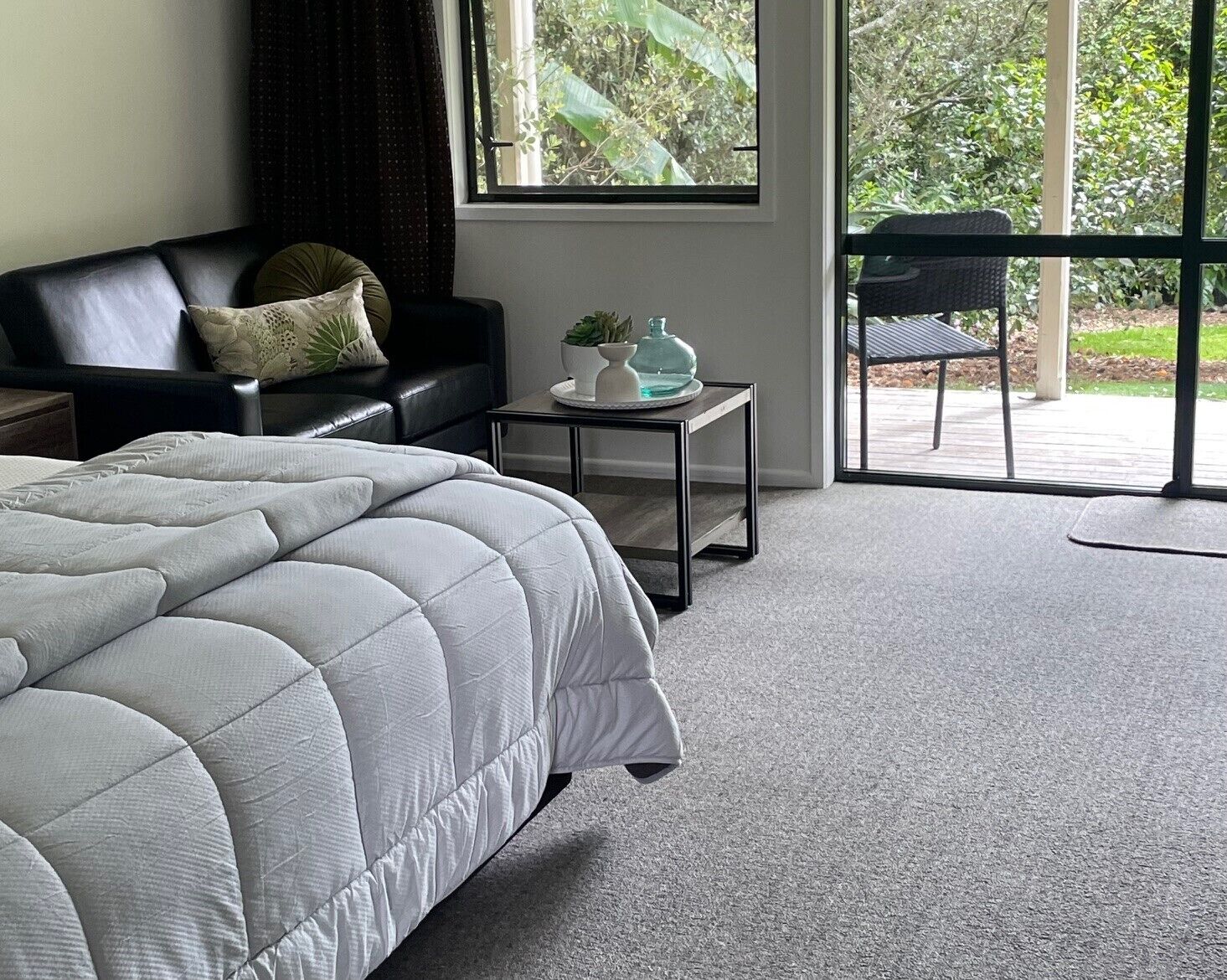 Ein Schlafzimmer mit einem großen Bett, einem schwarzen Ledersofa, einem kleinen Tisch mit Pflanze und Blick auf Grün durch das Fenster.