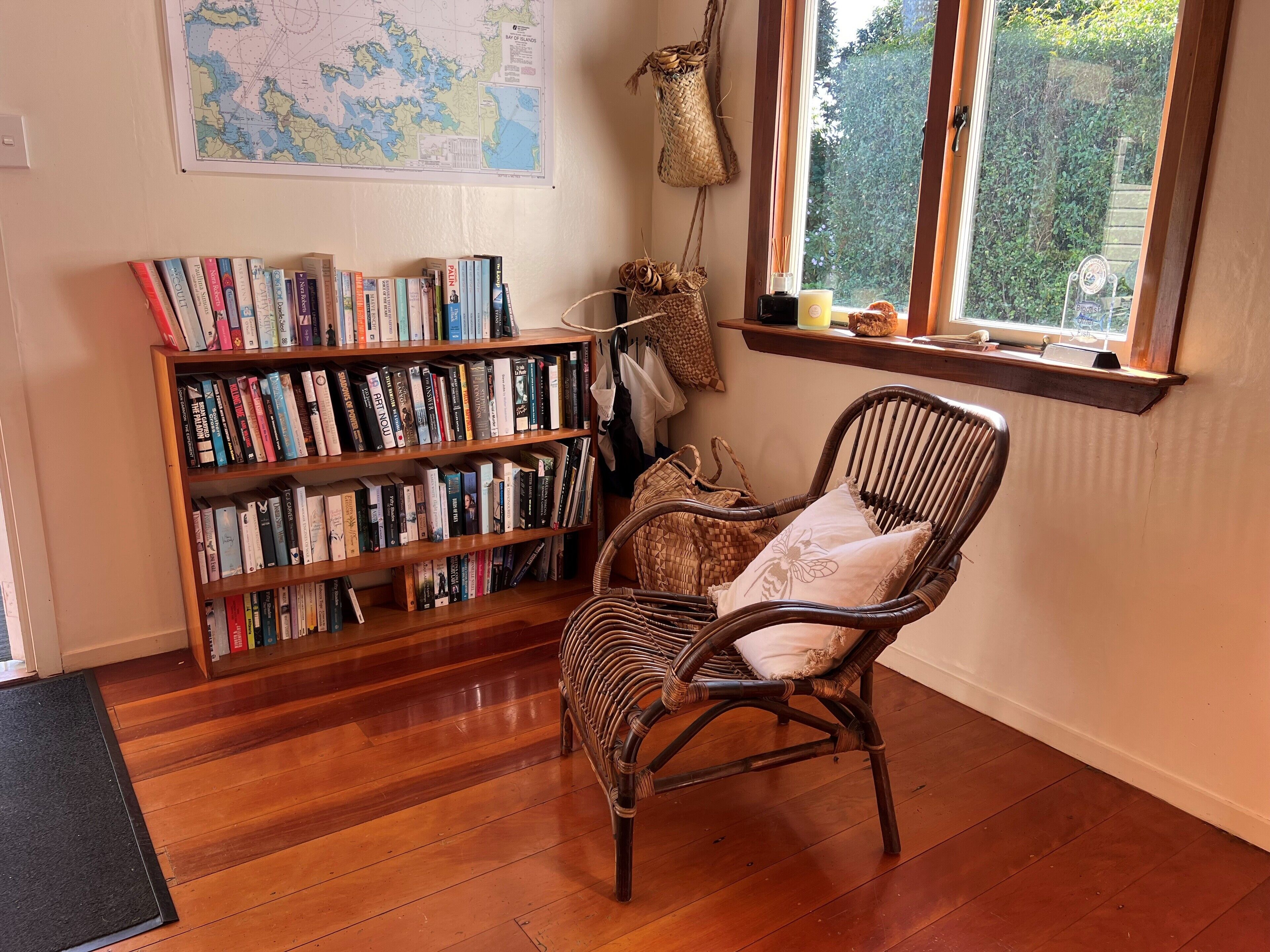 A cozy reading nook with a wicker chair, a bookshelf filled with books, a map on the wall, and a window with a view of greenery.