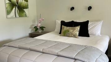 Una cama bien hecha con cubrecama acolchada, una sola almohada decorativa y una mesita de noche montada en la pared con un florero.