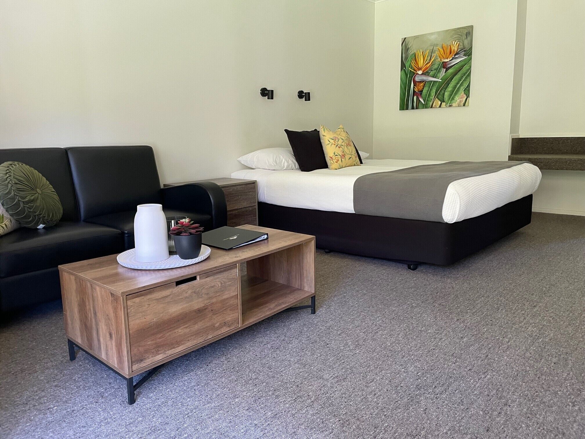 A modern hotel room with a black sofa, a wooden coffee table, a bed with a grey and white duvet, and a painting of a bird on the wall.
