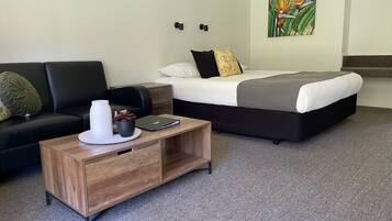 A modern hotel room with a black sofa, a wooden coffee table, a bed with a grey and white duvet, and a painting of a bird on the wall.