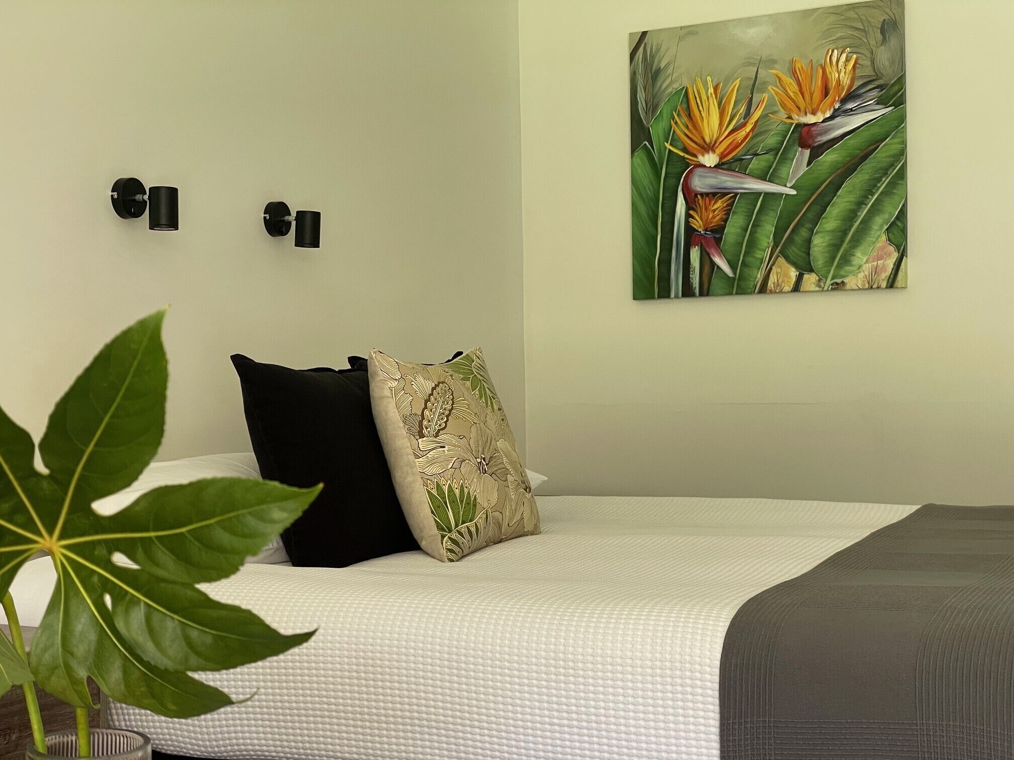 A bedroom with a bed, two wall-mounted light switches, a large painting of birds and foliage, and a potted plant.