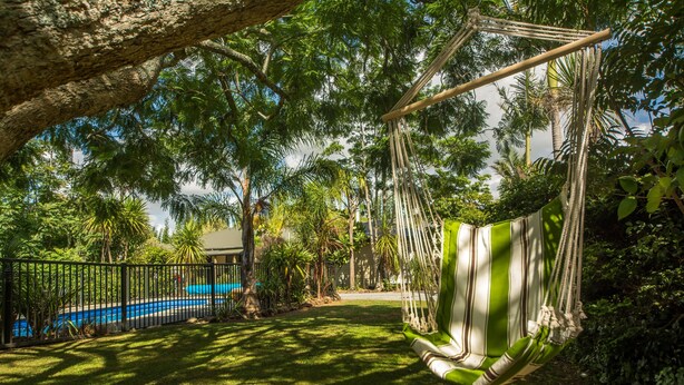A hammock swing in a lush garden with a pool and a fence. - Kauri Park Motel (Kerikeri)
