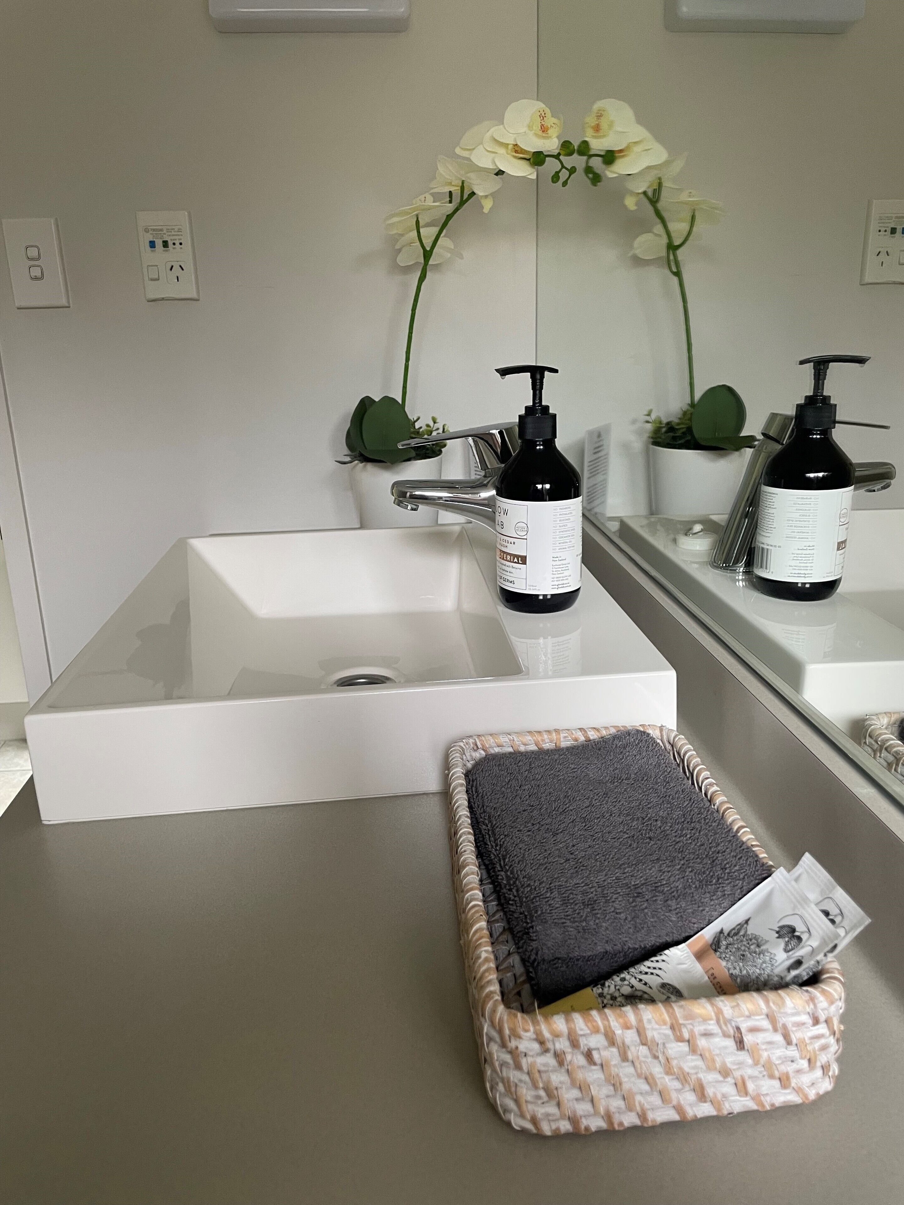 Un baño con un lavamanos blanco, dos dispensadores de jabón negros, un jarrón blanco con flores blancas, y una cesta tejida con una toalla negra.