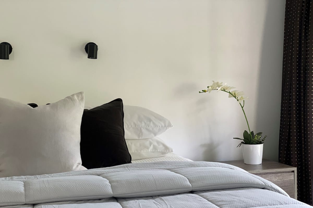 A neatly made bed with white and black pillows, a white textured duvet, and a potted orchid on a bedside table.