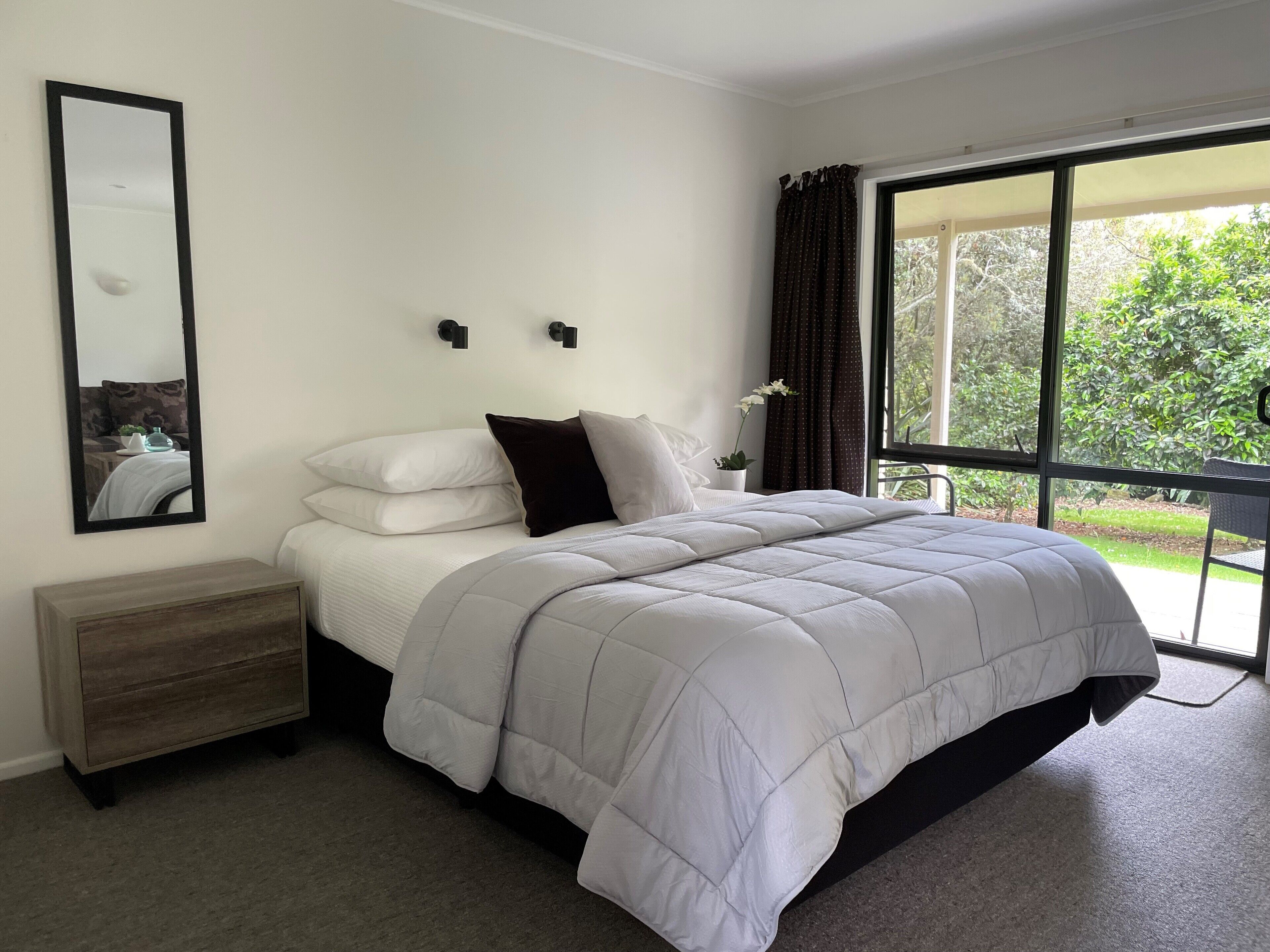 Une chambre avec un grand lit, une table de chevet, un miroir et une fenêtre donnant sur un paysage verdoyant.