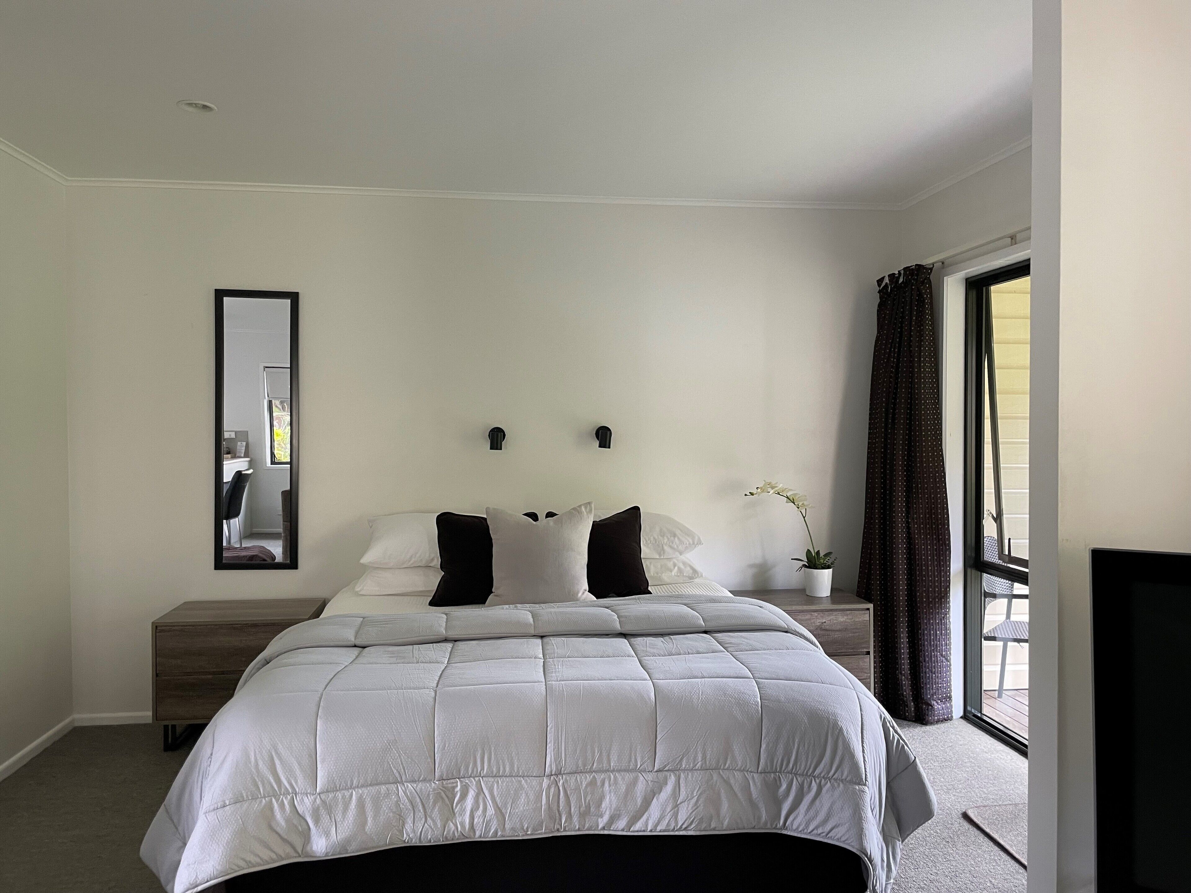A bedroom with a large bed, a bedside table, a mirror, and a window with curtains.