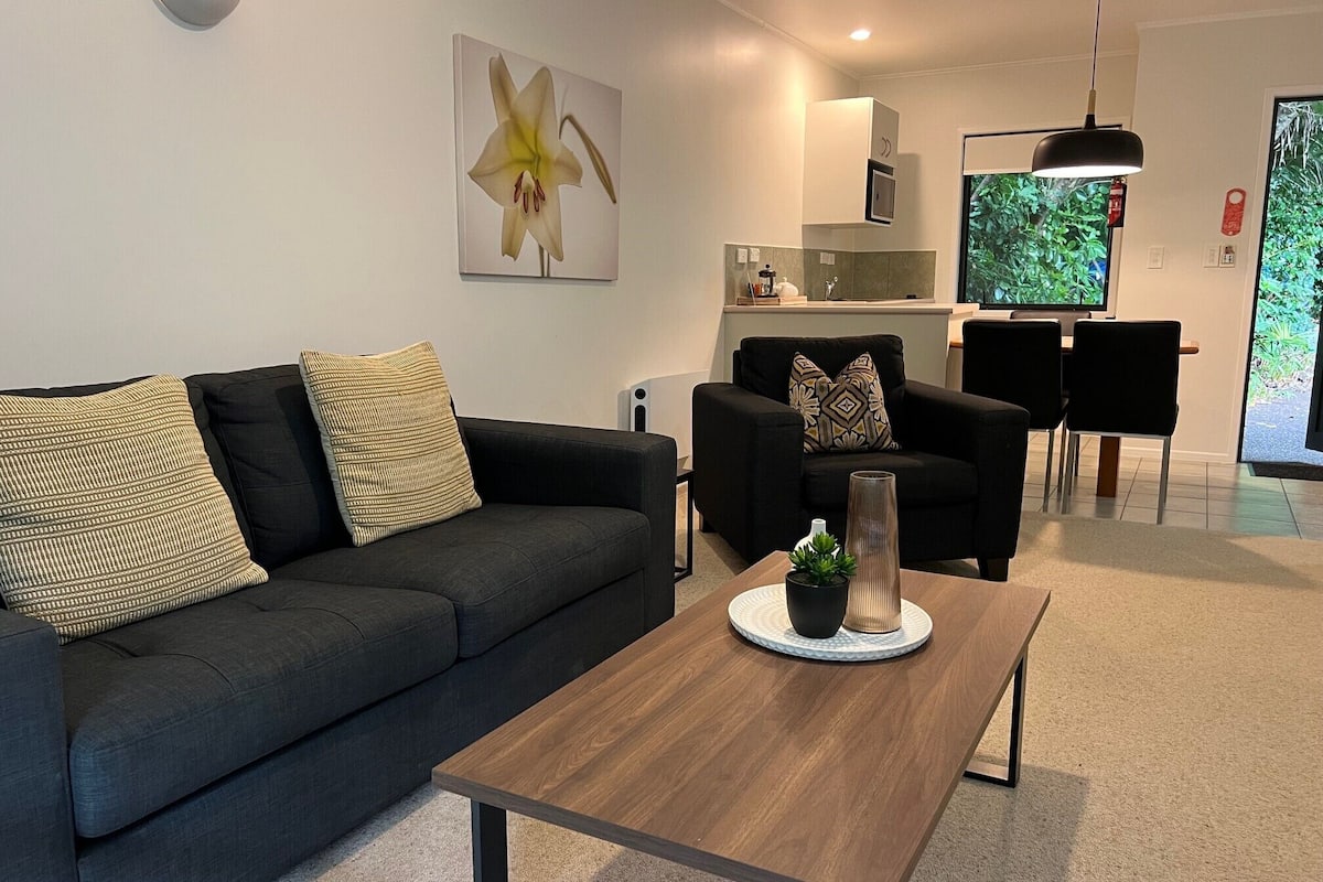 A modern living room with a dark grey sofa, a wooden coffee table, and a black armchair. There is a painting of a flower on the wall, a TV, and a dining area with a black table and chairs.