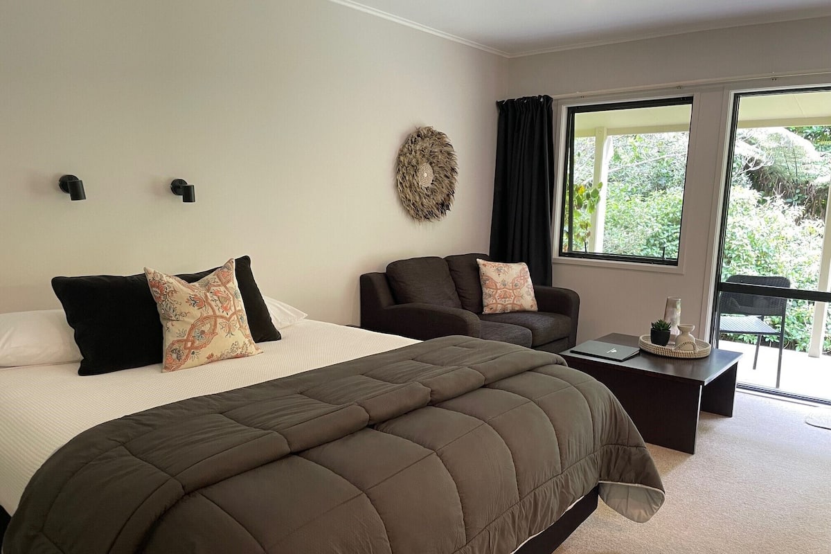 A bedroom with a large bed, a sofa, a small table, and a view of greenery outside.