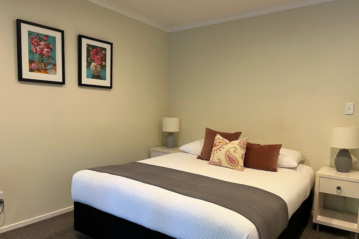 A bedroom with a bed, two bedside tables, a nightstand, and two framed pictures on the wall.