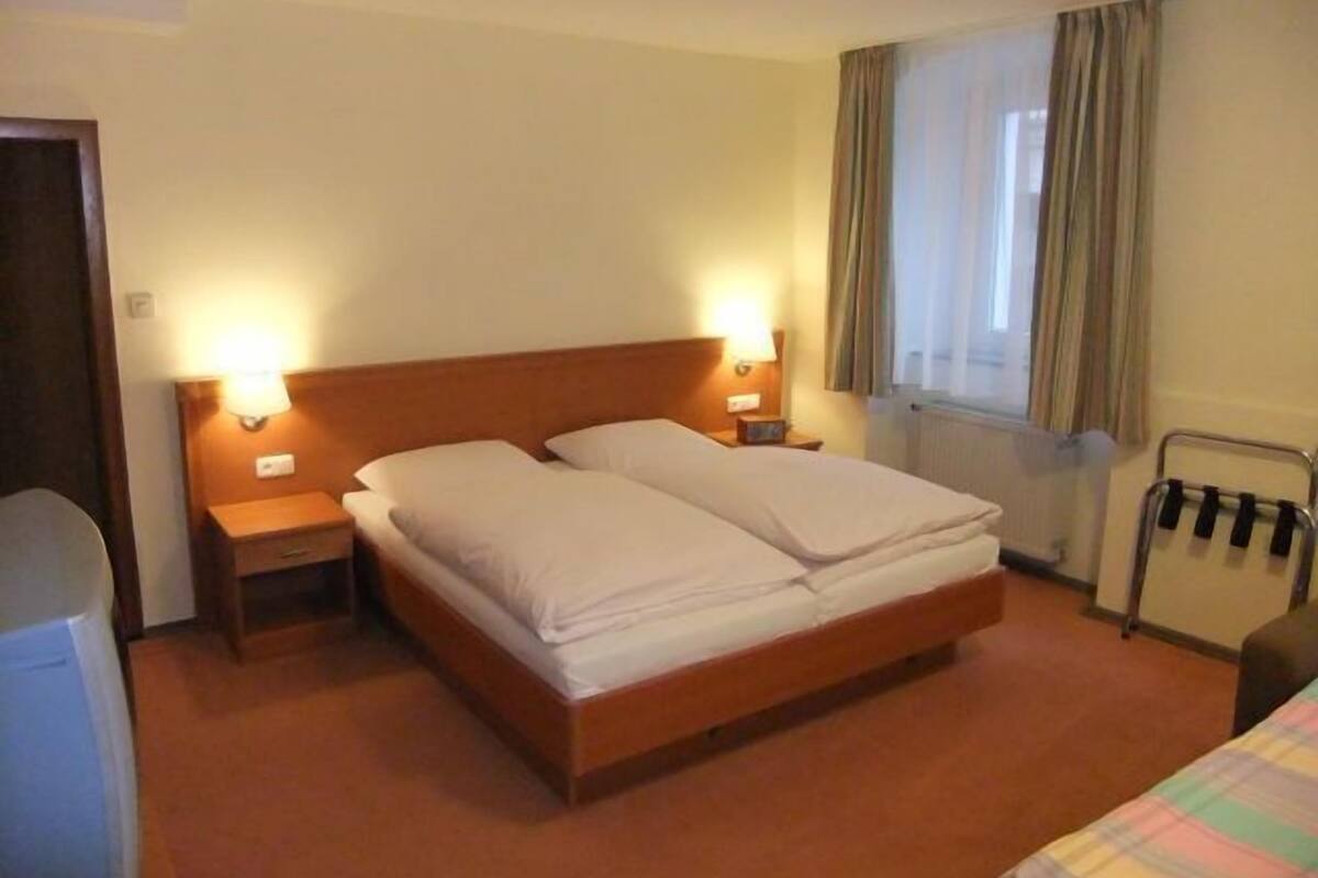 Habitación de hotel con una cama de madera, dos mesitas de noche con lámparas, un radiador y una ventana con cortinas.