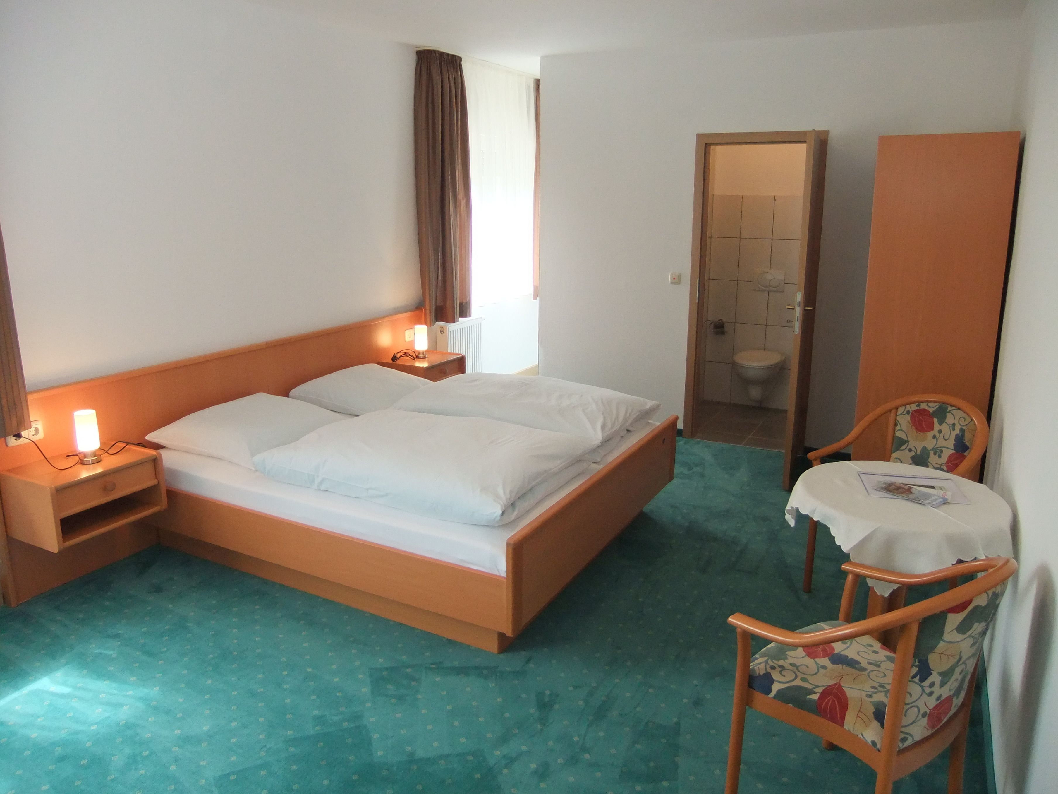 A hotel room with a bed, bedside tables, a chair, a small table, and a bathroom visible through an open door.