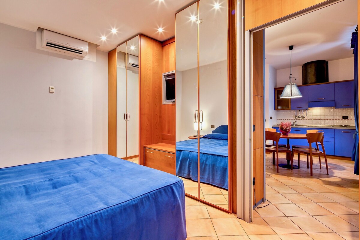 A bedroom with two beds, a wooden wardrobe, a small window, and a bathroom visible through a glass door.