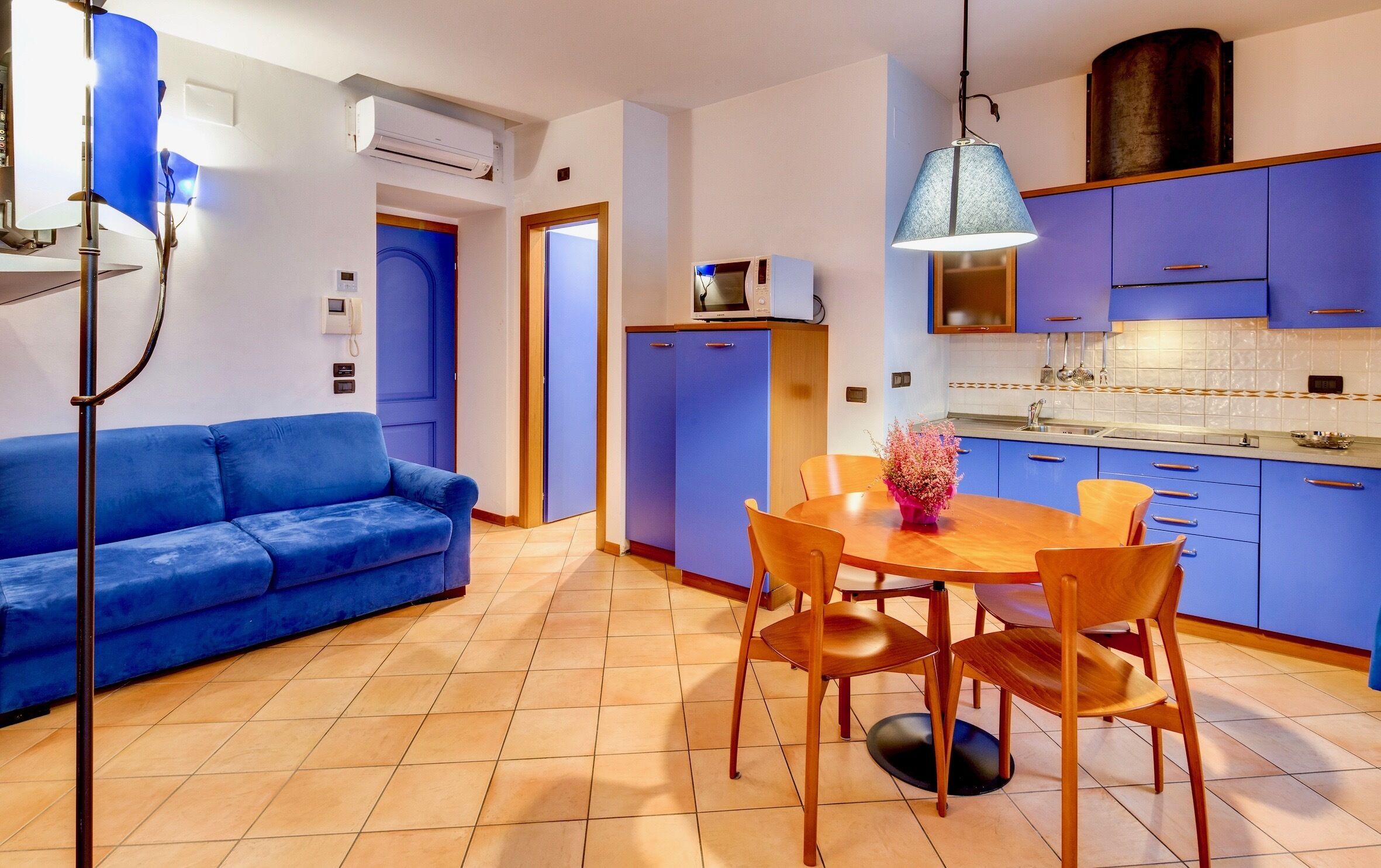 A compact living space with a blue sofa, a dining table with chairs, a kitchen area with blue cabinets, and a small refrigerator.