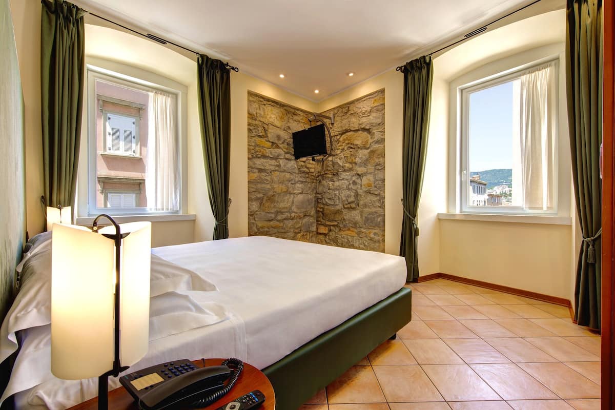 Une chambre d’hôtel avec un lit, une lampe, un téléphone, un mur en pierre et un téléviseur fixé au mur.